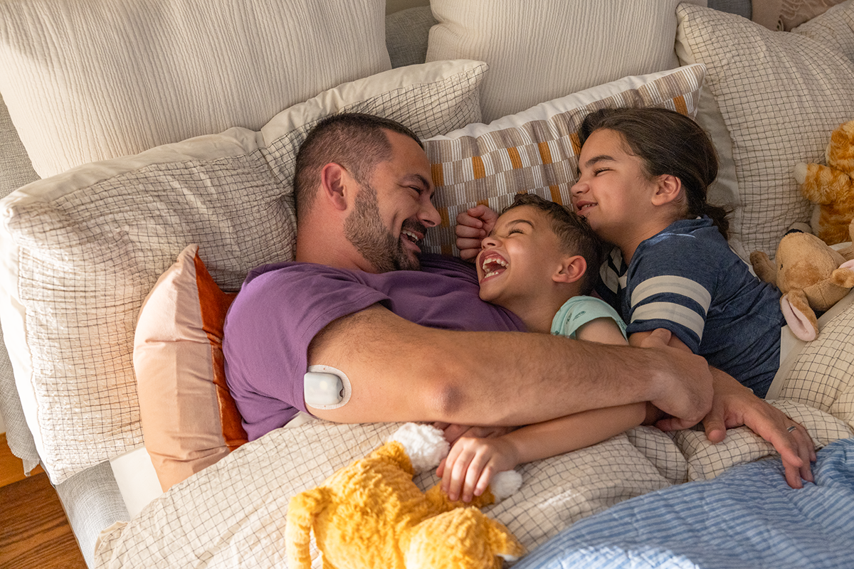 Father and two kids pileup in bed laughing with wither having an Omnipod Pod on his right arm