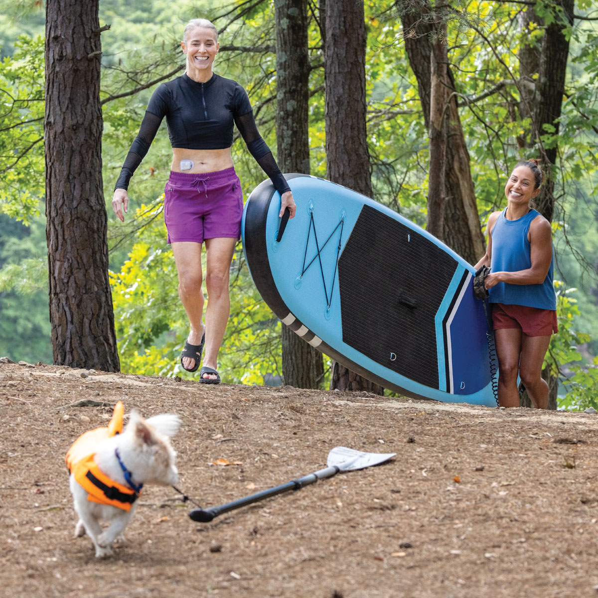 two people carrying a paddleboard with a dog looking on in front of them