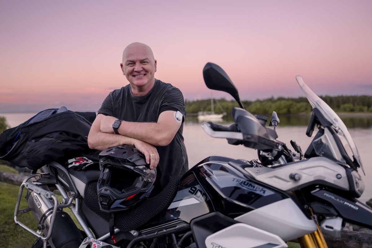 Scott, sitting on a motor bike, in the background is a pink sky and trees