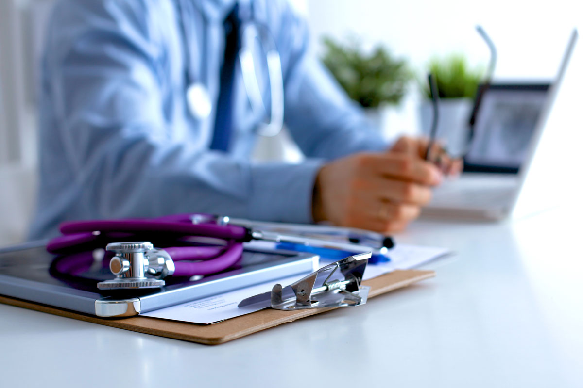 doctor sitting at desk holding glasses with a stethoscope on top of a clipboard