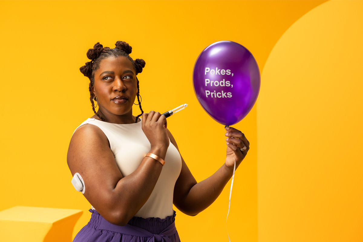 Woman wearing a pod on her arm holding a needle to a balloon that says Pokes, Prods, Pricks
