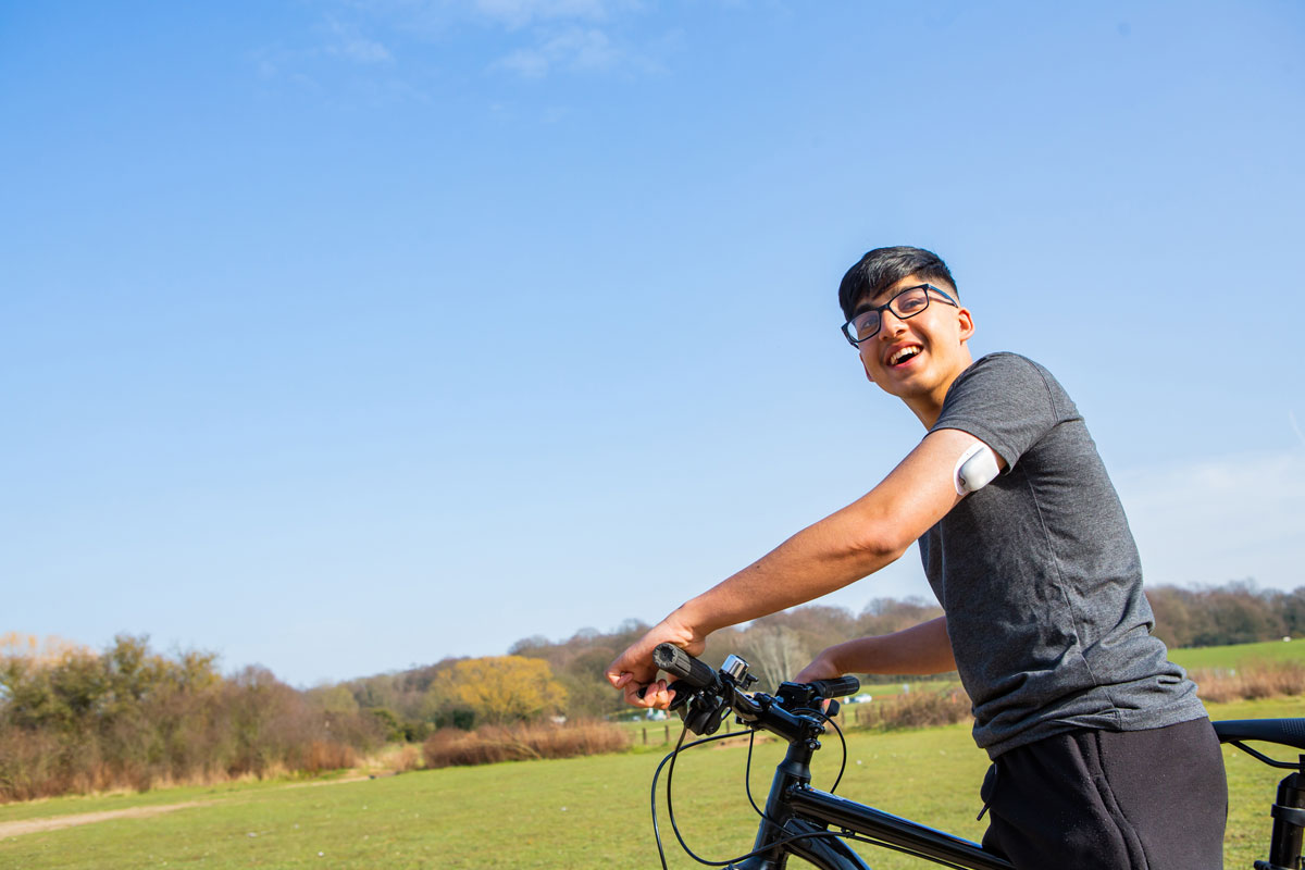 Dark haired young man with a Pod on the back of his arm smiling while standing next to a bicycle outdoors.