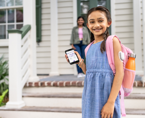 Una niña sonriente con una mochila, de pie delante de una casa con su smartphone.